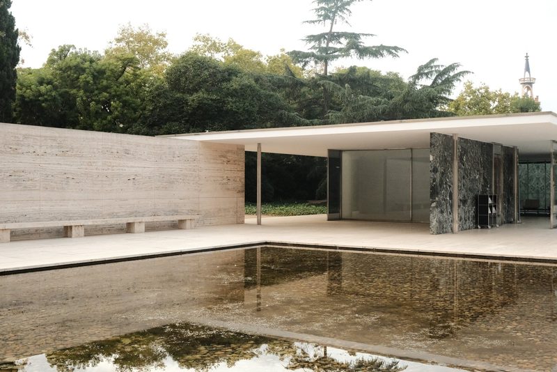 A modern building with a flat roof and a pool in front, featuring a wall with a bench and a row of trees in the background.