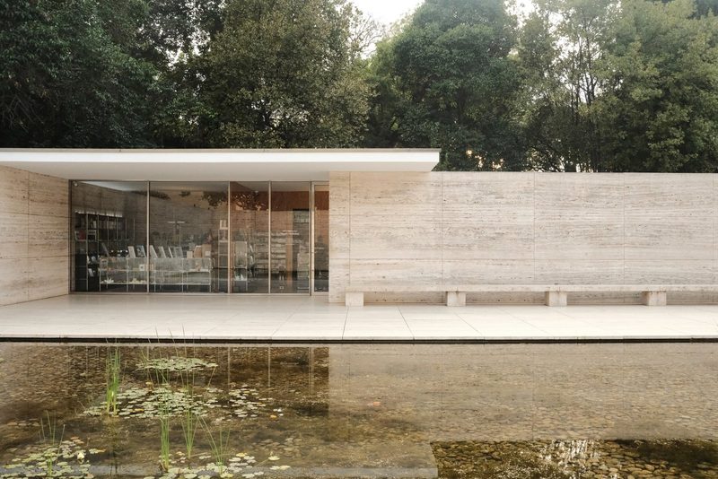 A modern building with a flat roof and large glass windows, featuring a wall and a pond in front.