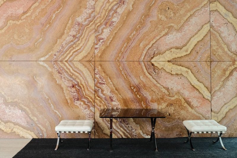 A table and two benches sit on a black rug in front of a large, marbled wall.