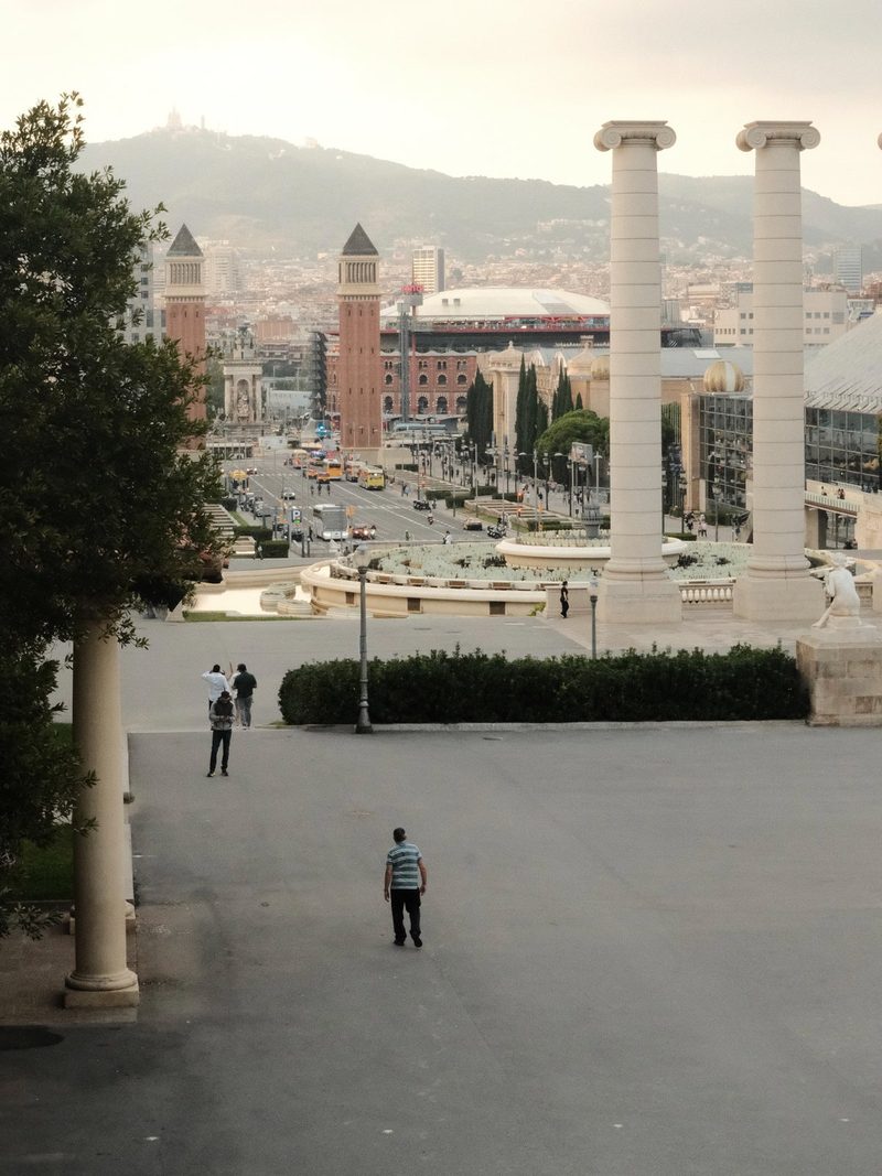 A cityscape with a large plaza, featuring a row of tall white columns, a large tree, and several people walking or standing in the foreground.