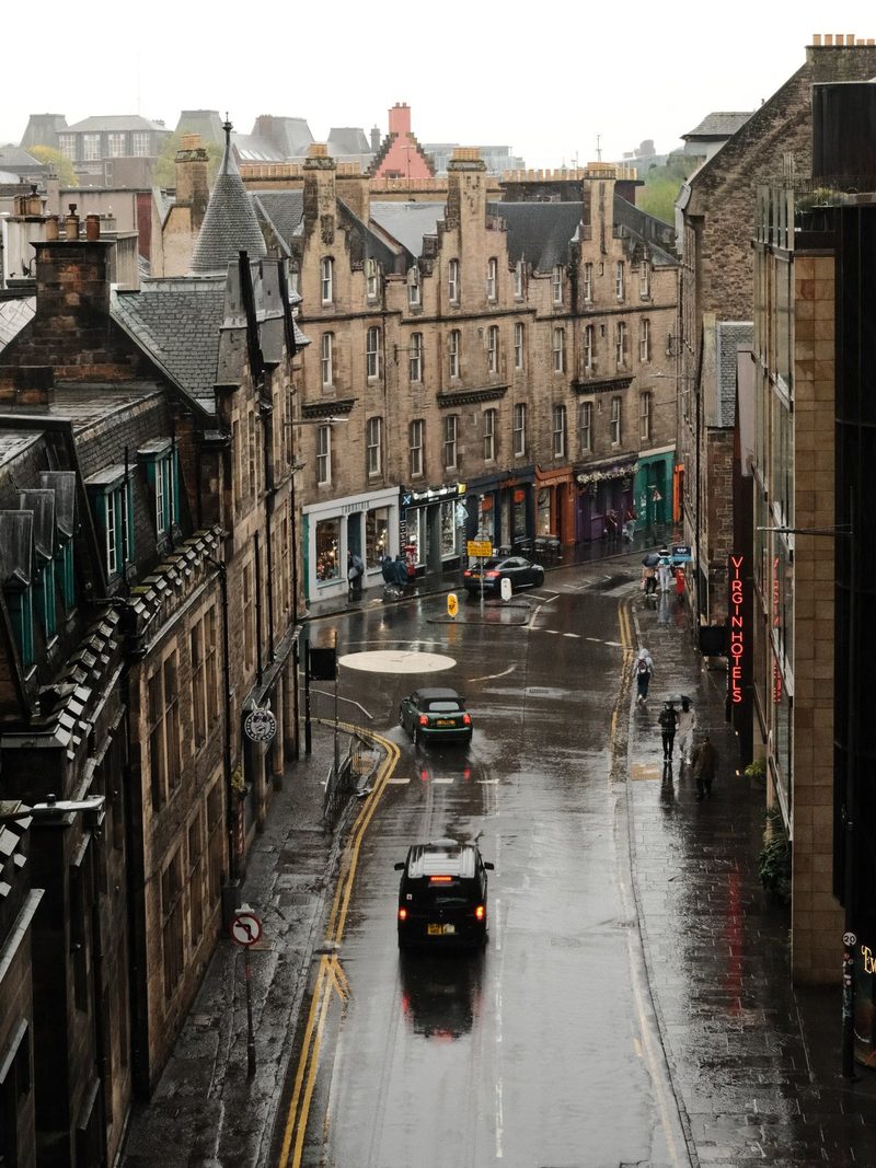 A city street lined with tall, multi-story buildings, featuring a mix of brown and tan hues, with a few cars and people visible.