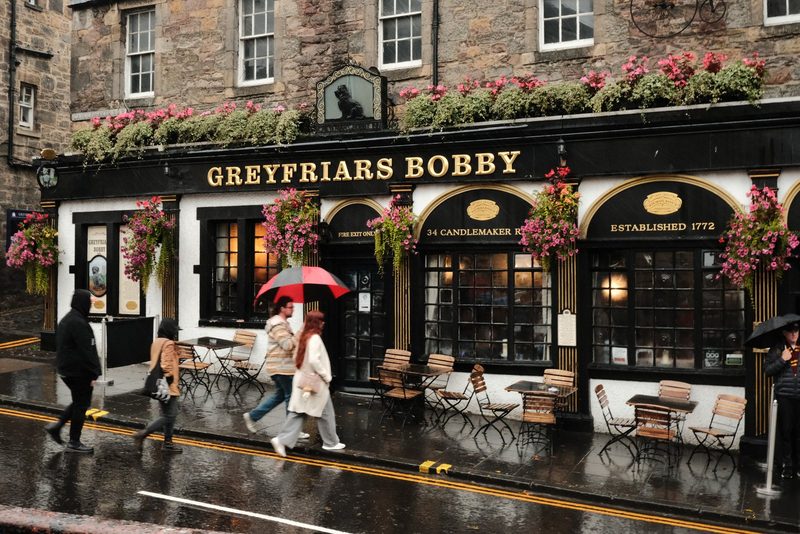 The image shows a storefront with a sign that reads "GREYFRIARS BOBBY" in gold letters, featuring a black and white facade with a row of tables and chairs outside.