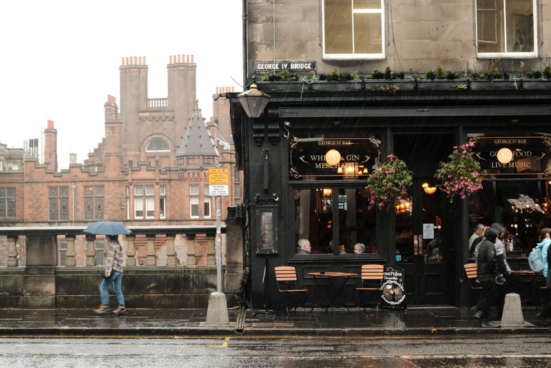 A street scene with a building featuring a sign reading "George IV Bridge" and a person walking with an umbrella.