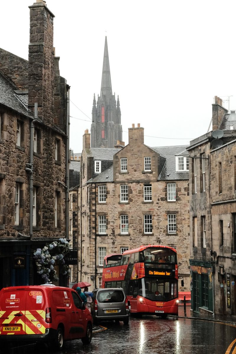 A city street scene featuring two red double-decker buses, a red van, and a grey car, with a tall stone building in the background.