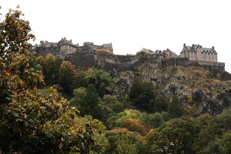 A large stone structure with multiple buildings and towers sits atop a rocky hill, surrounded by trees.