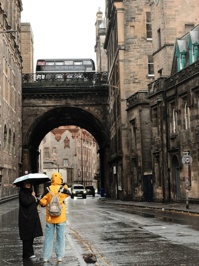 Two people, one wearing a yellow raincoat and the other in black, stand on a wet street, with a bridge and a bus in the background.