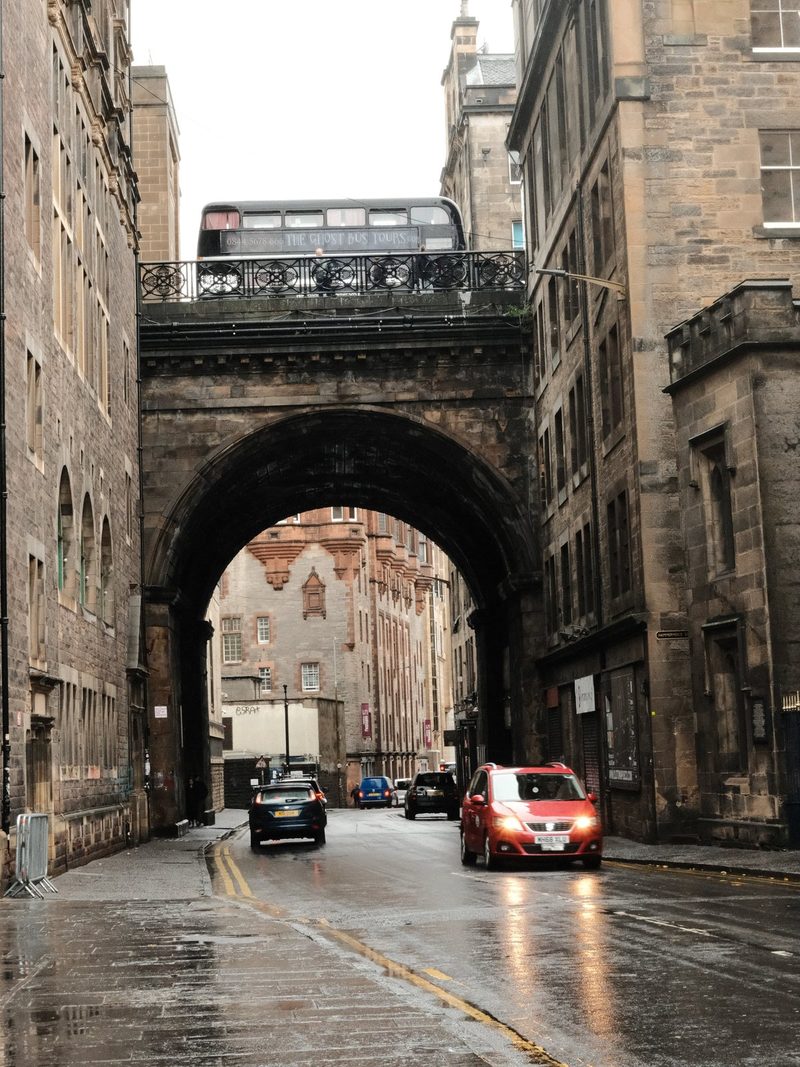 A stone archway spans a narrow street, with a black bus crossing it, and a red car driving below.
