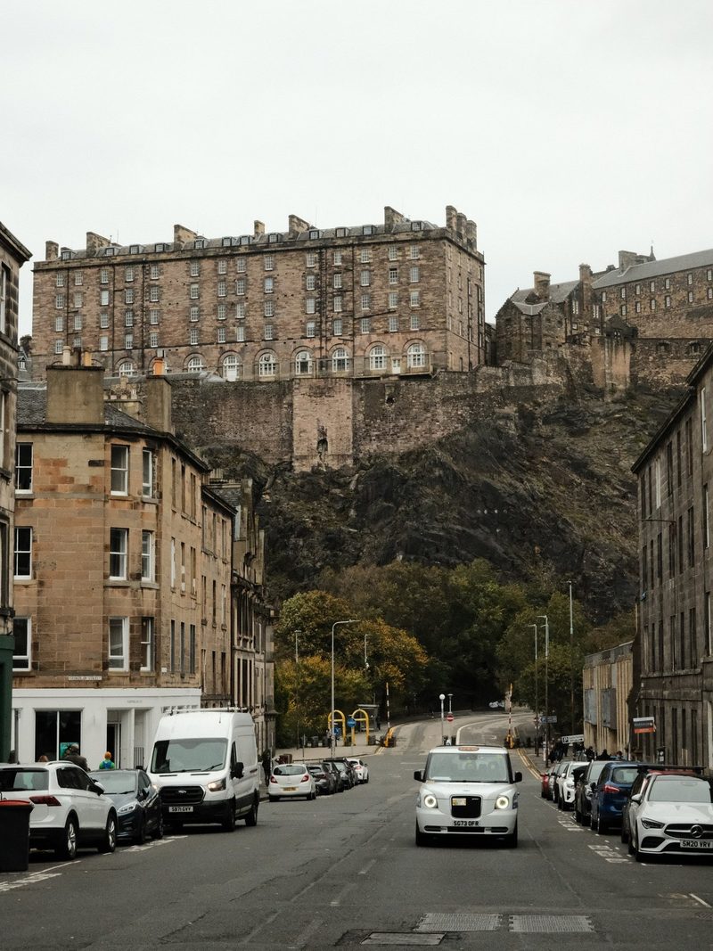 A large stone building with many windows sits atop a hill, surrounded by a row of smaller buildings and a street with parked cars.