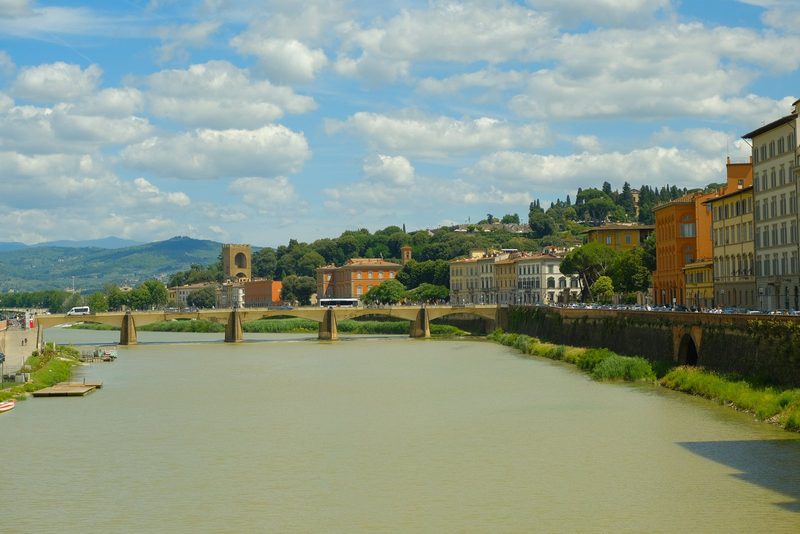 A river flows through a city, lined with buildings and a bridge, under a blue sky with white clouds.