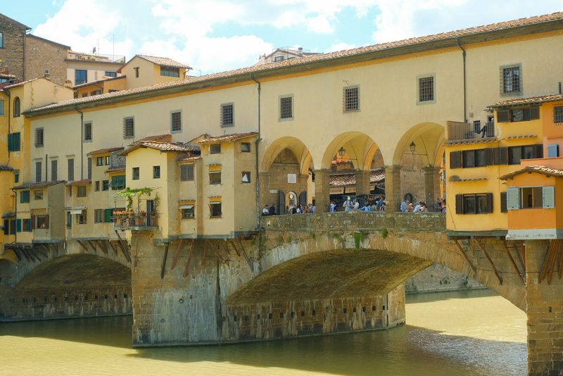 A stone bridge with arches spans a river, supported by a row of buildings on its side.