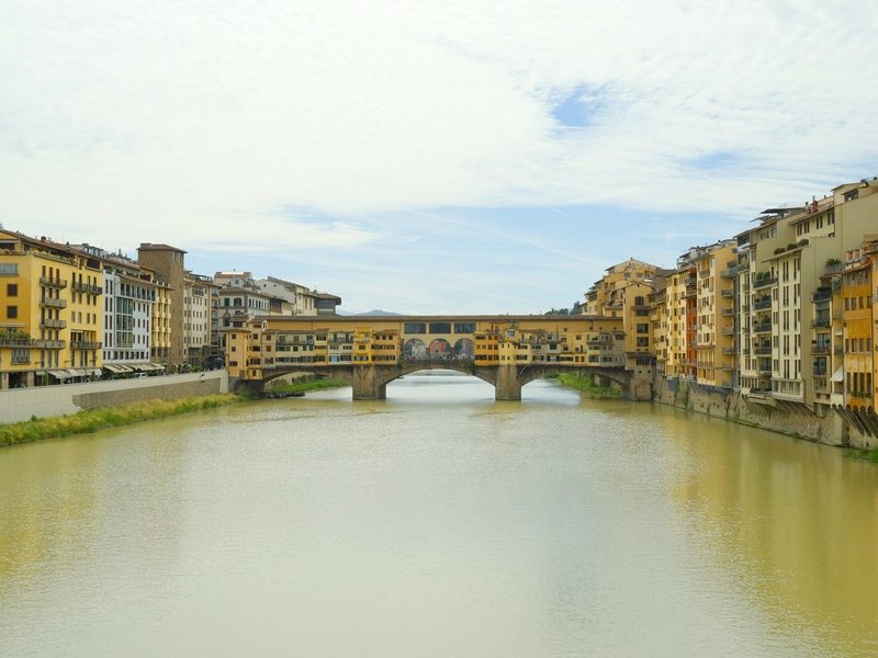 A stone bridge with arches spans a river, flanked by buildings on either side.