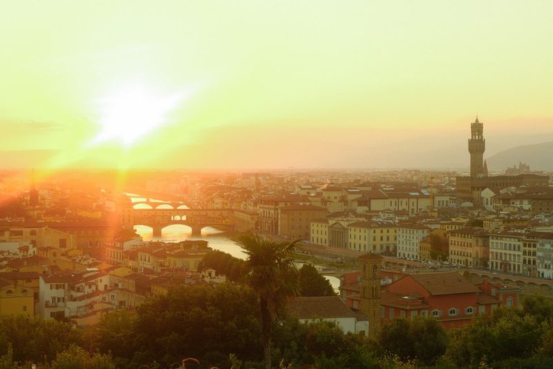 A cityscape with a river running through it, featuring a bridge and a tall tower, set against a hazy sky with the sun shining brightly.