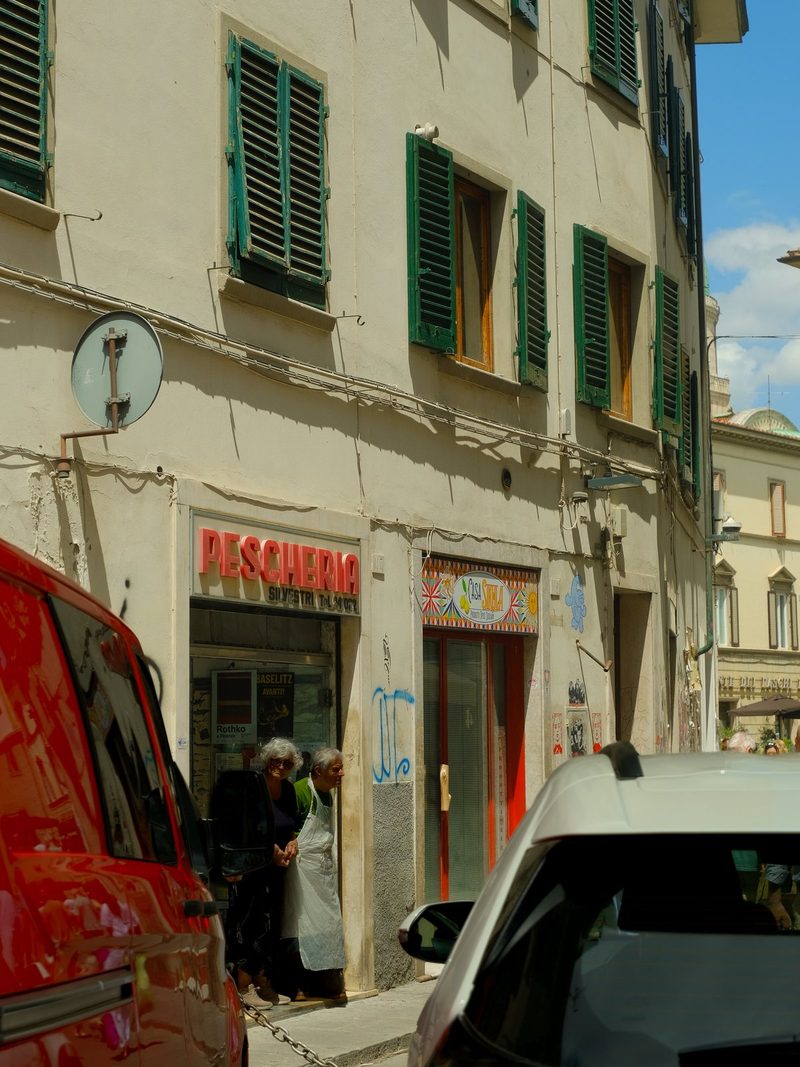 Two elderly women, dressed in white aprons, stand in the doorway of a shop, with a red van parked in front of them.