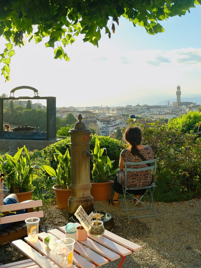 A woman sits on a chair, facing a cityscape with a water fountain and a table with drinks in the foreground.