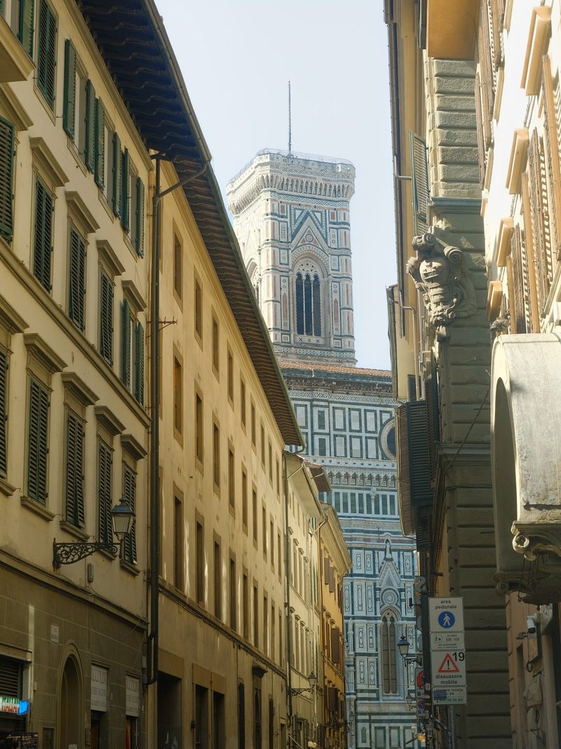 A tall, ornate building with a tower and a clock face, situated between two rows of buildings with windows and shutters.
