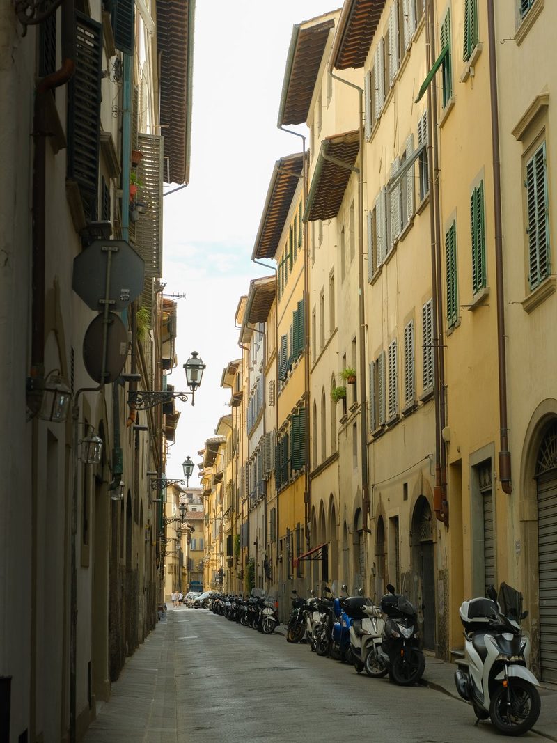 A narrow street lined with tall, yellow buildings, featuring green shutters and a row of parked motorcycles.