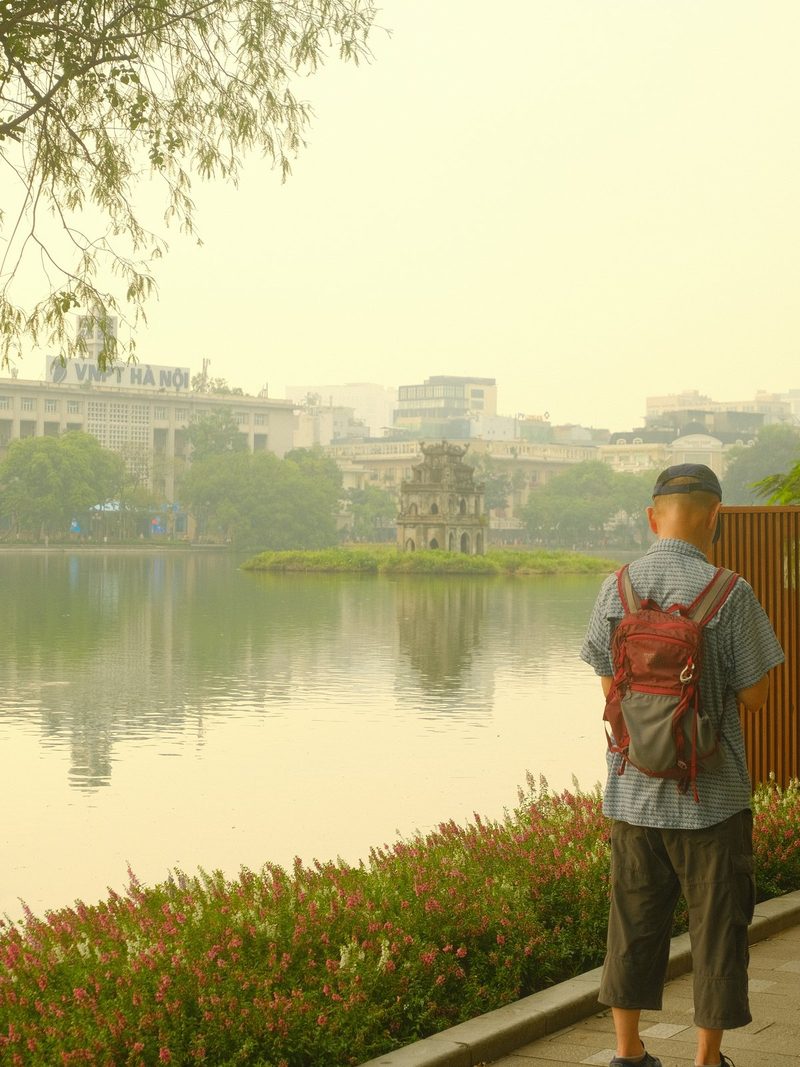 A man stands on a sidewalk overlooking a lake with a cityscape in the background.