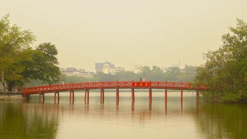A red bridge over a lake in a city with trees and buildings in the background.