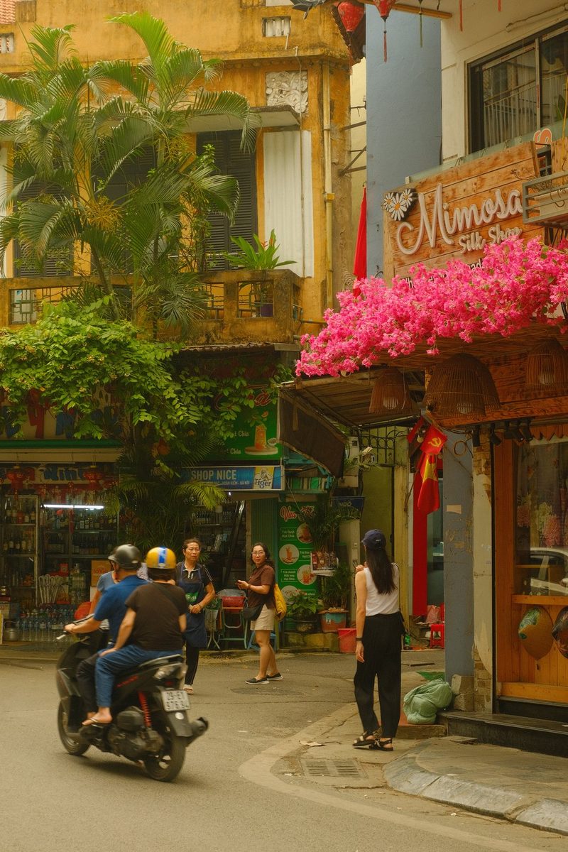 A street scene in a tropical city with a mix of old and new architecture, featuring a prominent sign for a silk shop and a group of people on a scooter.