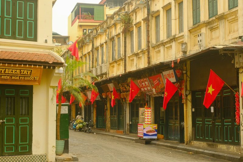 A street in a city with a mix of old and new buildings, featuring red flags and a motorcycle.