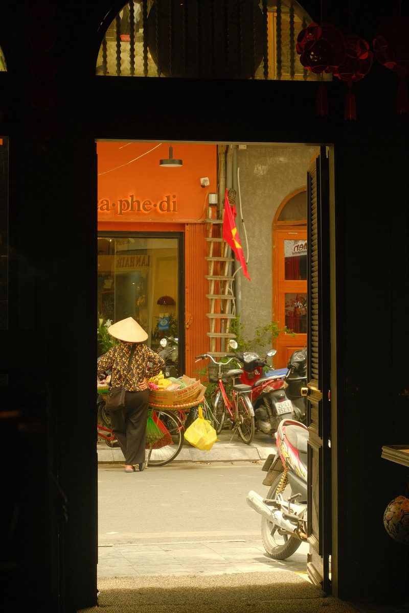 A street vendor in a traditional Vietnamese hat sells goods in front of a shop with a red facade and a ladder leaning against the wall.