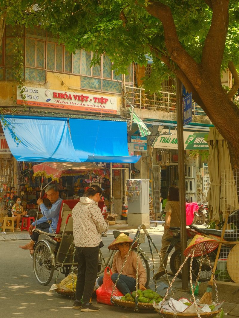 A street scene in a Vietnamese city, with a tree, a man on a tricycle, and a woman selling fruit.