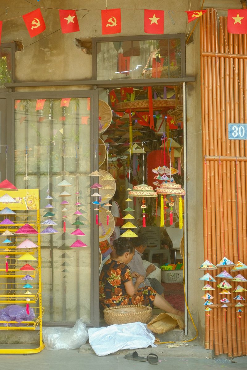 A storefront in Vietnam sells traditional conical hats, with a woman sitting outside sewing one.