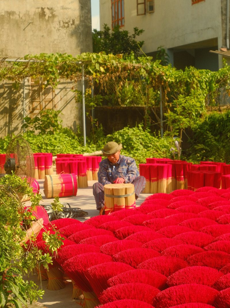 A man is sitting in a courtyard surrounded by rows of red and pink bundles of fabric, possibly preparing for a textile production process.