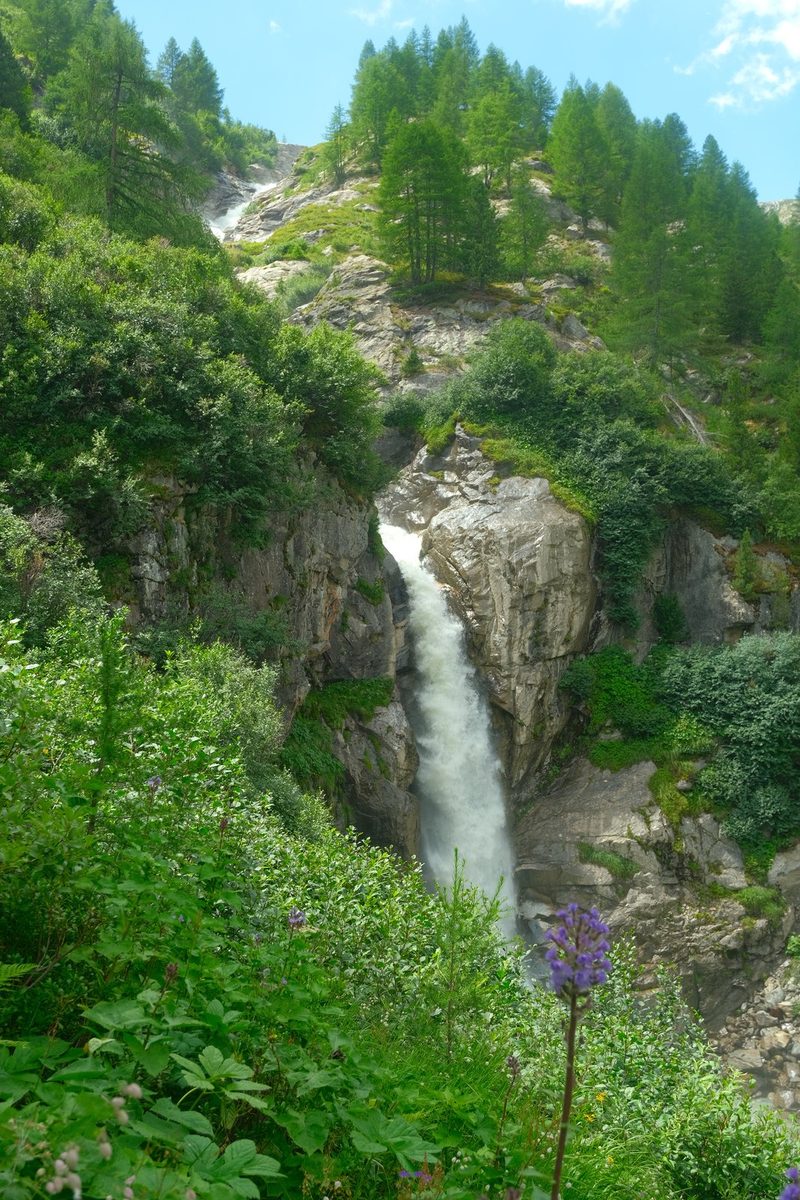 A waterfall in a mountainous area surrounded by trees and greenery.