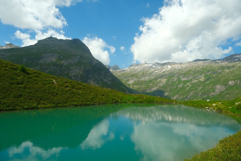 A serene mountain lake surrounded by lush greenery and majestic mountains under a blue sky with white clouds.