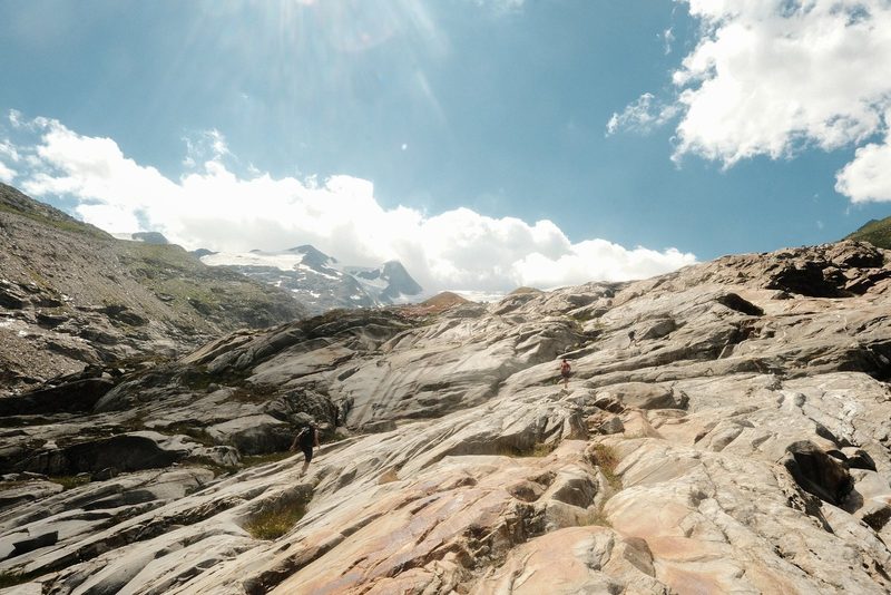Two people hiking on a rocky mountain trail under a blue sky with clouds.