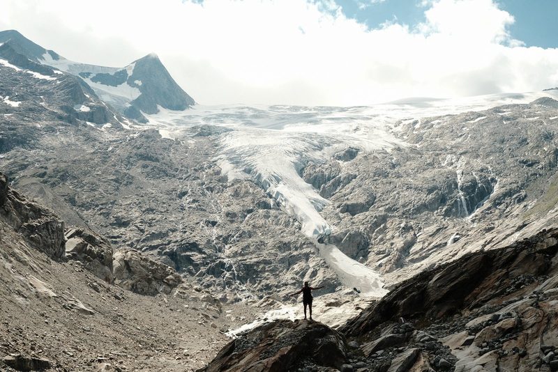 A person stands on a rock in front of a large glacier in a mountainous area.