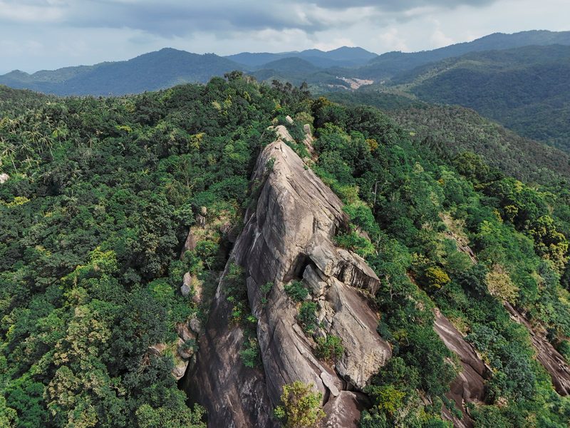 A large, light-colored rock formation is surrounded by lush greenery, with a mountainous landscape in the background.