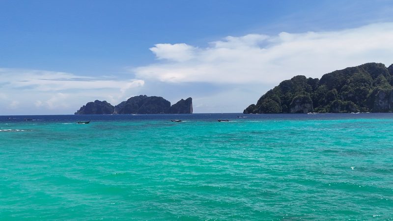 A large body of water with a few boats and a rocky island in the distance, under a blue sky with clouds.