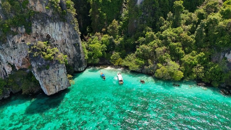 Three boats float in a turquoise sea, surrounded by steep cliffs covered in lush greenery.