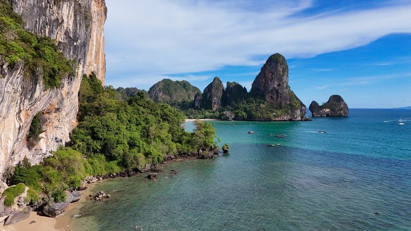 A rocky cliff rises from a sandy beach, with a body of water in the foreground and a distant island with similar cliffs.