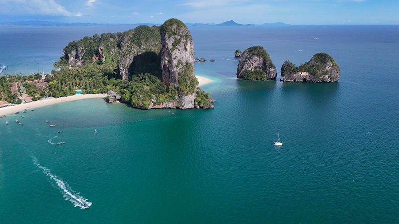 A large body of water with a sandy beach and several rocky islands, featuring a few boats and a clear blue sky.