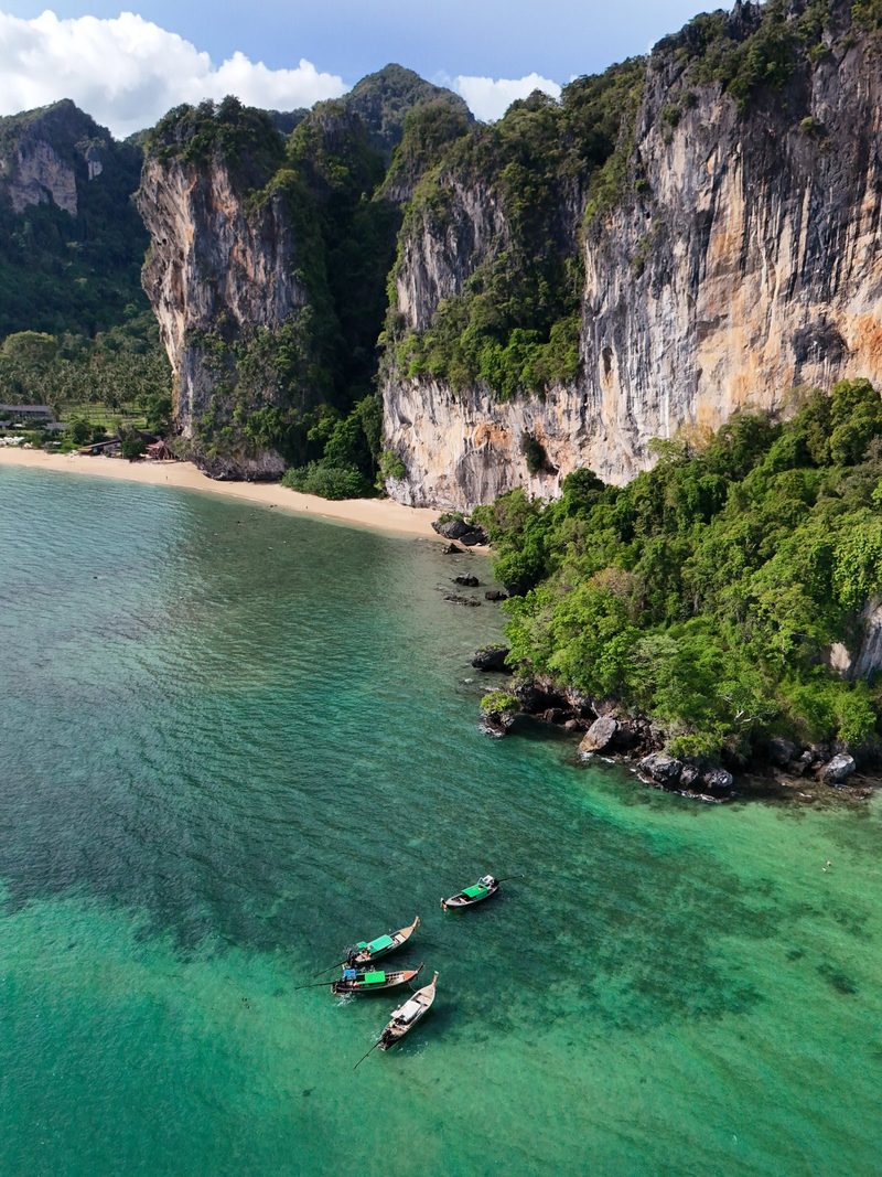 A group of small boats float in the shallow, clear waters of a tropical bay, surrounded by steep cliffs covered in lush greenery.