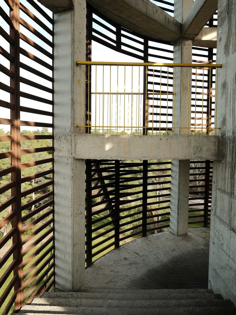 A concrete structure with a yellow railing and wooden slats, featuring a staircase and a view of trees and a road.