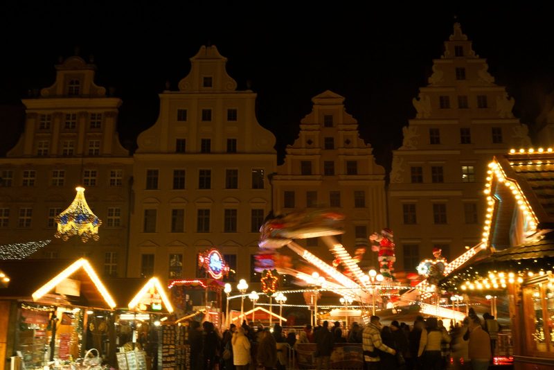A large, multi-story building with a distinctive roofline stands behind a vibrant, illuminated carnival or amusement park, featuring a variety of rides and attractions.