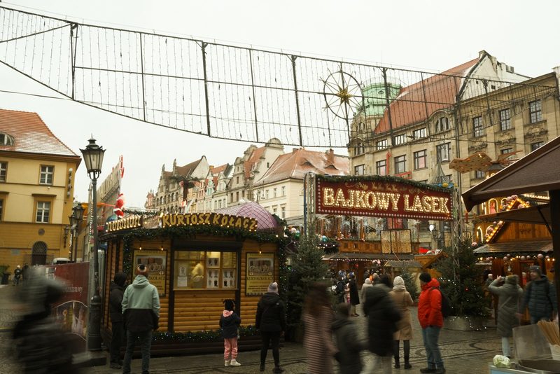 A festive outdoor market scene featuring a wooden building with a sign reading 'KURTOSZ KOLACZ' and a red sign with white lettering reading 'BAJKOWY LASEK'.