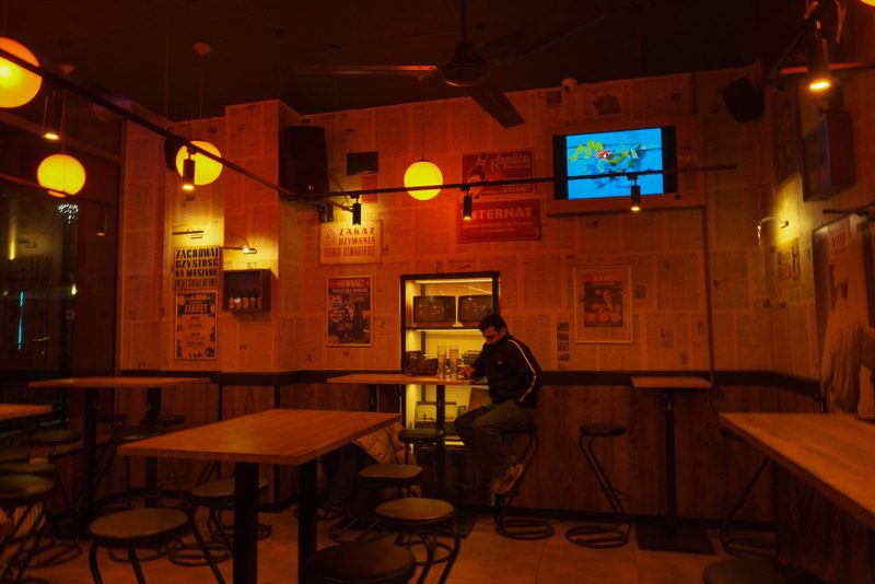 A man sits at a bar in a dimly lit room, surrounded by tables and chairs, with a television on the wall and a shelf displaying bottles and glasses.