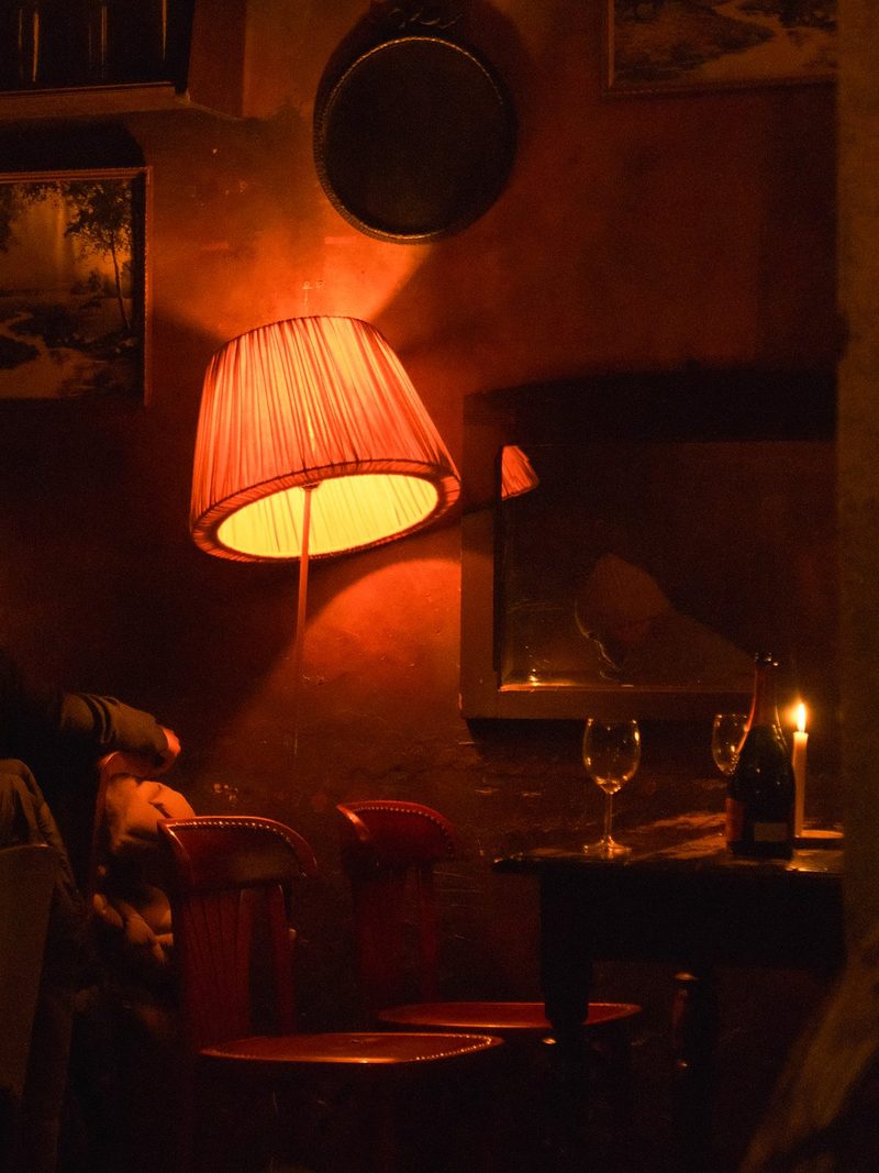 A dimly lit room with a lamp, a table with wine glasses, and a chair, featuring a mirror and a painting on the wall.
