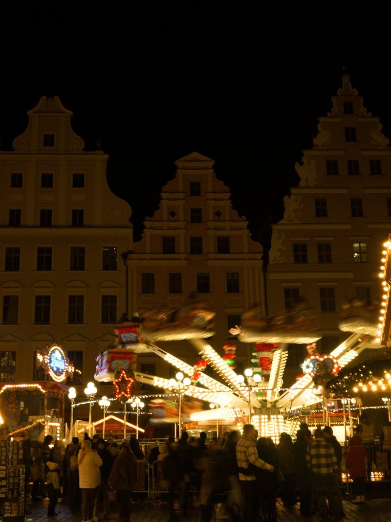 A large, multi-level, ornate building with many windows and a distinctive roofline stands behind a brightly lit, spinning ride with people in the foreground.