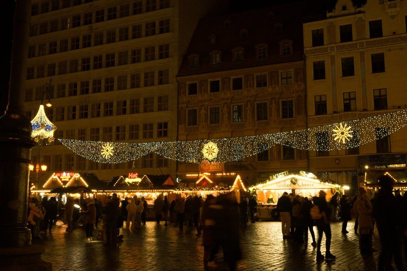 A large, open square is filled with people and decorated with festive lights and snowflake-shaped decorations.