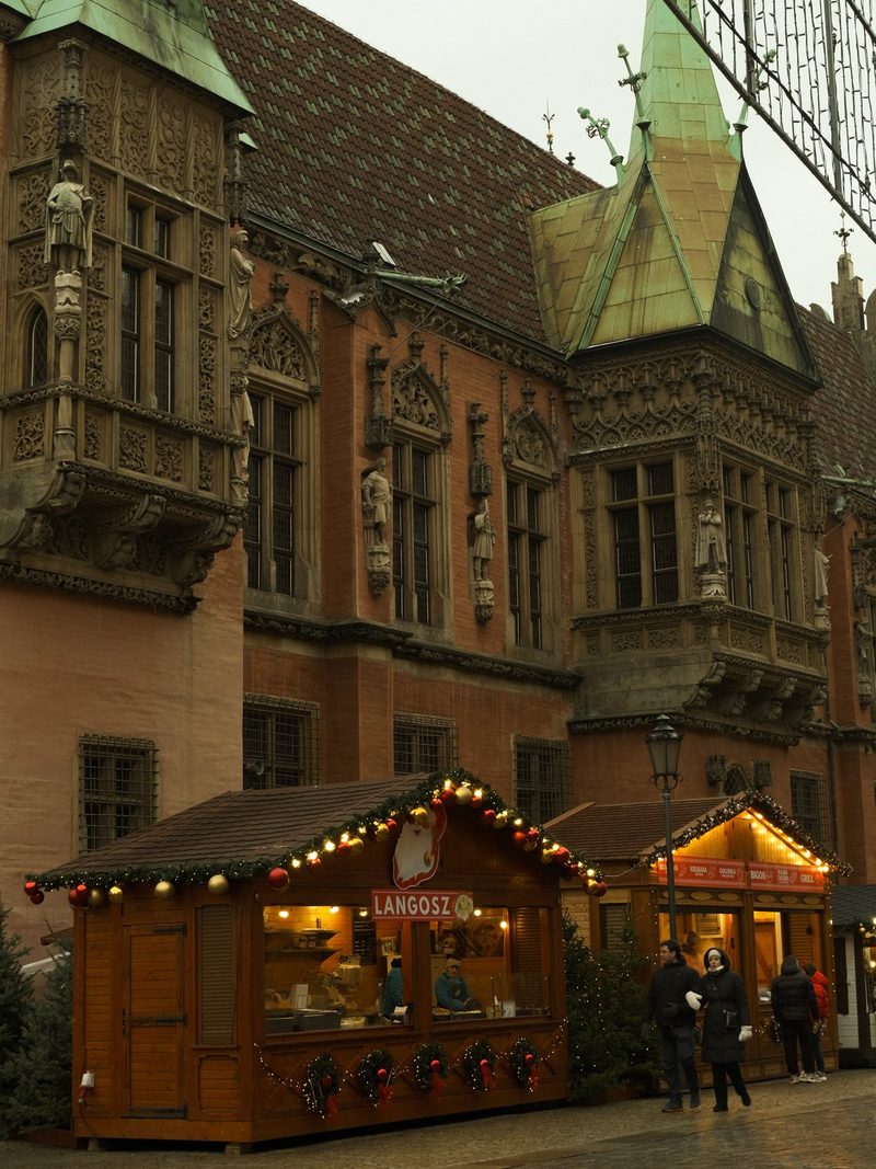 A large building with a red brick facade and ornate stone carvings, featuring a row of wooden stalls with festive decorations and signs in a foreign language.