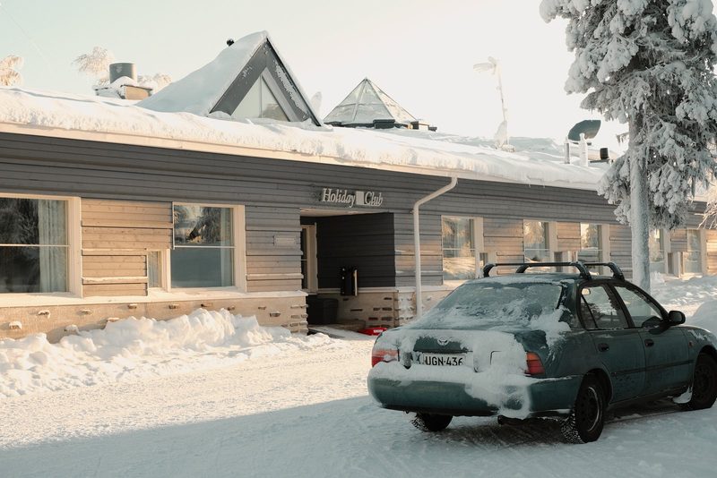 A car is parked in front of a building with a sign that reads "Holiday Club". The car is covered in snow, and the building has a snowy roof.