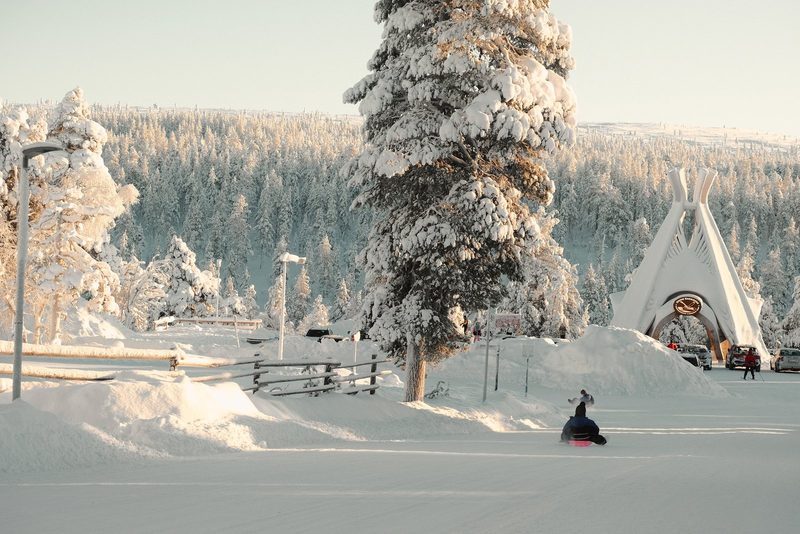 A person on a snowmobile is driving away from a large white structure with a circular opening, surrounded by snow-covered trees and a snowy landscape.