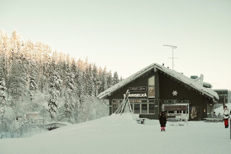 A dark-colored building with a sloping roof and a sign that reads "LIEGGA LAAVU" and "SARISELKA" is situated in a snowy landscape, surrounded by trees and people.
