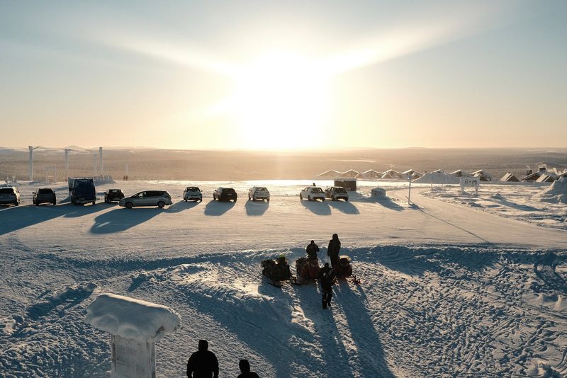 A group of people in winter clothing stand in a snowy landscape, with a row of parked cars in the background.